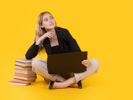 Student girl with laptop and books. Girl is sitting cross-legged on the floor. Girl leans against a stack of books. Student is sitting on the floor with laptop. Isolated on orange. Training, educationの写真素材