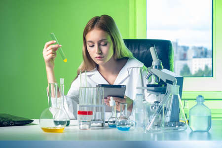 Chemistry researcher works in laboratory. Girl compares the results of the experiment with the control data in the tablet. Laboratory assistant at work. A woman with a flask and an electronic tablet.の写真素材