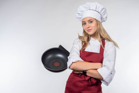 Beautiful blonde woman chef holding pan. Chef girl isolated on white. Female cook in white uniform and hat keeping frying pan. Cooking and food concept - smiling female chef or cook with pan and spoonの写真素材
