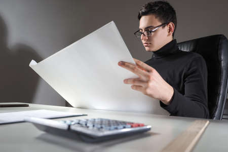 Auditor at work. Calculator next to man. Concept - business audit. Male auditor thinks something. He provides audit services for businesses. Young guy is studying documentation. Auditor career.の写真素材