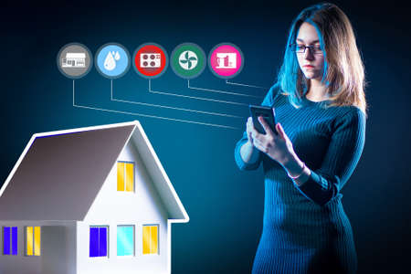 Woman controls smart home. Layout of a house next to a businesswoman. IOT icons next to girl. She sets up a smart home using her phone. Application for setting up a smart home system. IOT applictionの写真素材