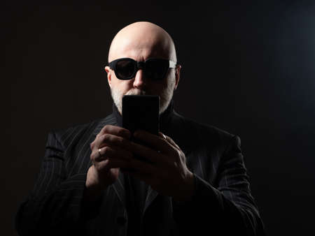 Man holds a smartphone in front of him. Concept - he makes a video call or a selfie. Portrait of a businessman on a dark background. Man makes a video call via phone. Bald businessman in sunglassesの写真素材