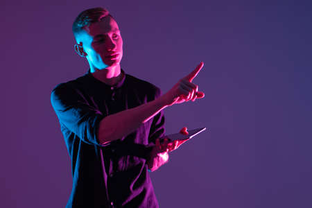 Young guy reaches out to screen. Concept - he clicks on a virtual screen. Portrait of a young man with a tablet in his hands. Student dressed in casual style. Student with tablet on purple backgroundの写真素材