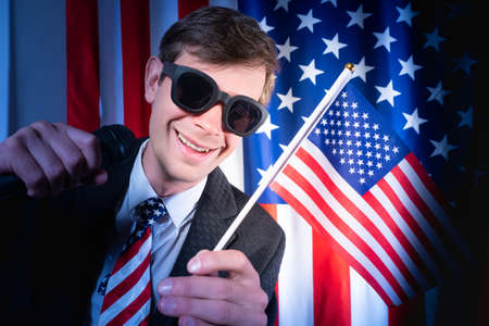 American showman. Showman with microphone on background of USA flag. American with a microphone in his hands. Career in USA show business. Radio host from United States. Young guy in sunglasses.の写真素材