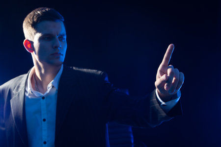 Business man pushing imaginary button. Young business man in classic suit. Human on a dark background. He reaches out to screen. Guy clicks on virtual screen. Man stretches his finger to monitorの写真素材