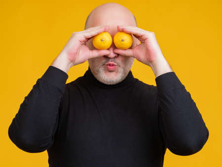 Man with lemons in his hands. He shows lemons instead of eyes. Grown man is grimacing. Portrait of a human on a yellow background. Man puts lemons to his eyes. Concept - use of citrus fruits.の写真素材