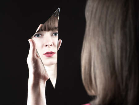Woman with a shard of a mirror in her hands. Her face is reflected in broken mirror. Concept - female depression. Midlife crisis in women. Girl suffers from psychological depression. Black backgroundの写真素材