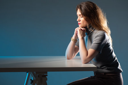 Woman praying with closed eyes. Girl praying while sitting at table. Catholic girl during prayer. Catholic or Orthodox turns to God. She sleeps sitting at table. Woman crossed her arms under her chinの写真素材