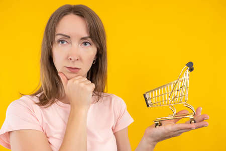 Purchases, consumer basket. A thoughtful woman with a miniature shopping cart. Purchasing power. Reduced purchasing power. Supermarket cart as symbol of shopping. Orange background.の写真素材