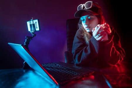 Blogger woman next to a laptop. Girl holds a cup of tea in her hands. She records video on her smartphone. Blogger woman broadcasts video. Woman student with laptop in a dark room. Online streamingの写真素材