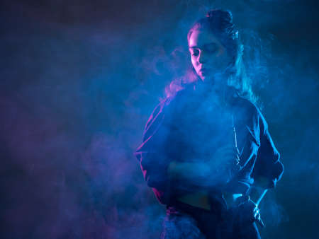 Vape pen in hands woman. Portrait of a girl in clouds of cigarette smoke Young woman smokes electronic cigarette. Vaper is holding a vape pen. Student with a vaping gadget. Vaper girl in neon lightの写真素材