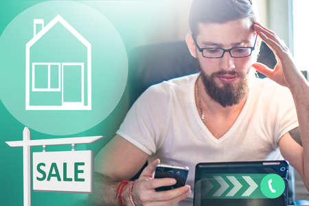 The realtor advises clients online. Sale of real estate. A man sells a house. Buying a house. A man with a smartphone and a tablet next to a sign about the sale of a house.の写真素材