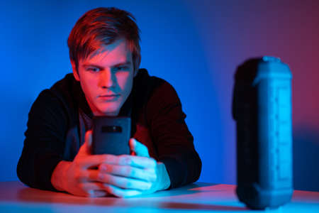 Intelligent speaker next to a person. He controls portable speaker through phone. Partret of a young guy with a smartphone in his hands. Black intelligent speaker in foreground. Buy black columnの写真素材