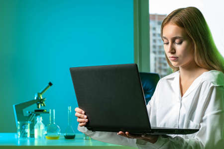 A girl with a laptop on the background of a chemical laboratory. Experiments and research. Chemical tests. Lab assistant with laptop. A woman in a white coat. Laboratory tests.の写真素材