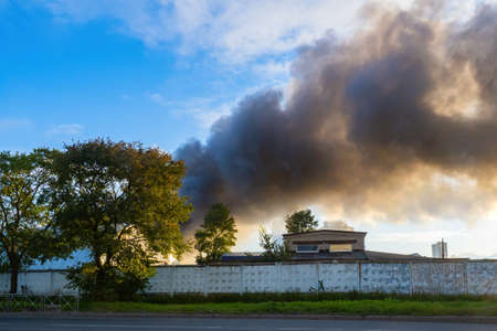 Fire fume in an industrial zone. Smoke behind a stone fence. Building behind the fence is on fire. Smoke from the fire against the blue sky. The factory is on fire. A fire at an industrial enterpriseの写真素材