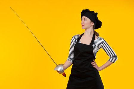 Chef woman. Cookie is holding a sword. Woman in chef uniform. Portrait of a female cook with a sword. Concept - she cuts something. Chef cuts something with a sword. Girl in black cook hat and apronの写真素材