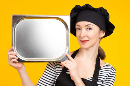 Portrait of chef kitchen. Woman chef kitchen with empty tray in hands. She smiles while holding gastronomic bowl. Chef kitchen on yellow background. Girl in a cook's uniform shows an empty trayの写真素材