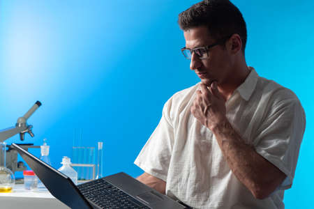 Young scientist on background of laboratory. Scientists with laptop are thinking about something. Concept career as scientist. Man in white shirt near microscope. He is engaged in scientific researchの写真素材
