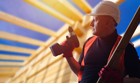 Builder during work. Blurred timber frame behind builder. Construction of wooden house. Man with a...