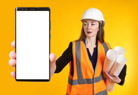 Female civil engineer with smartphone. Girl in construction helmet and reflective vest demonstrates...