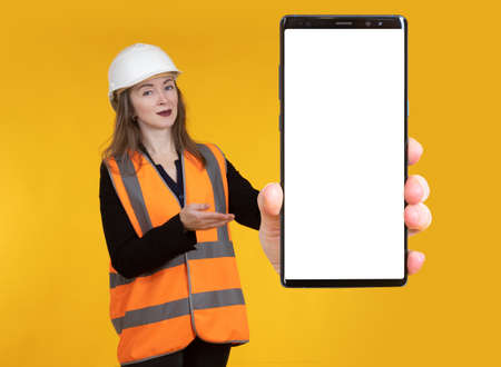 Calling a repairman via mobile app. Girl demonstrates smartphone with place for text on the screen. A woman in a construction helmet and a reflective vest. Advertising of a construction company.の写真素材