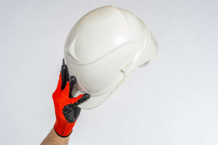 White construction helmet in hand. Close-up safety helmet. Man makes greeting gesture with construction helmet. Gloved hand raises casque above his head. Casque on light background.の写真素材