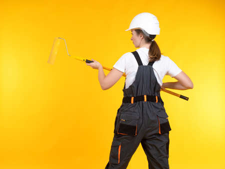 Female house painter. Woman stands with her back and paints the wall yellow. Girl with a painting tool. A woman in overalls and a hard hat. Repairwoman on a yellow background. Bright colors concept.の写真素材