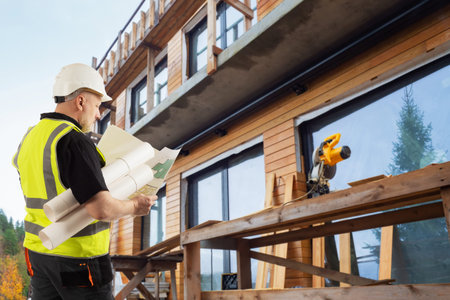 Builder next to a wooden cottage. Man examines drawings against the background of a house under...