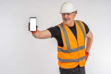 Cell phone hand builder. Foreman shows phone. Cellphone for advertising construction apps. Builder in yellow vest. Smartphone template with empty space. Place for text on phone screen. Happy builderの写真素材