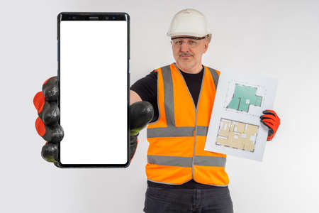 Architect shows empty phone. Builder hand with smartphone mockup. Place for text on screen. Smartphone copy space. Giant builder hand with cellphone. Man in builder's clothes on light backgroundの写真素材