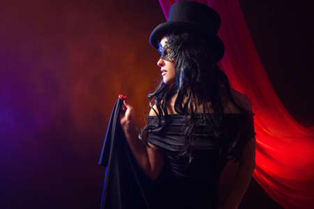 Girl in lace mask. Concept - a girl plays in the theater. Mask of lace. Clothing for a theme party. Woman gathered for a masquerade ball. Girl on a background of red cloth. Woman is all in blackの写真素材