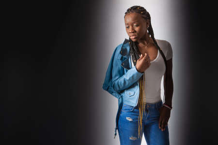 Dark-skinned girl near the wall. Woman with african pigtails. African demonstrates urban clothing. Model shows the outfit. Intimidated black woman. Sad look. Girl with dreadlocks on a dark backgroundの写真素材