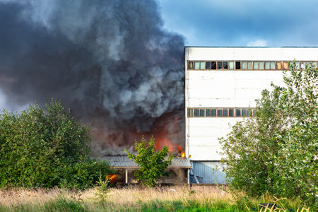 Fire next to industrial building. Black smoke from fire. Fire in factory or factory. Black smoke on blue sky background. Concept is industrial accident. Flame next to industrial building.の写真素材
