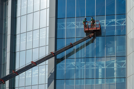 Workers are washing walls of building. Two men on boom of tower crane. Window cleaners at work. Workers wash glass skyscraper. Concept - building cleaning services. Window cleaning company employeesの写真素材