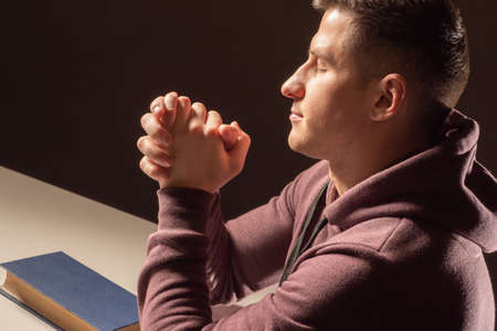 man is praying. Portrait of Prayer guy. Young guy is praying with his eyes closed. Christian folded his palms for prayer. Prayer man on dark background. Book with praying in front of Catholicの写真素材