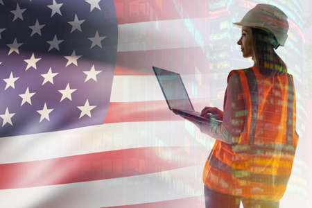 A female engineer in USA. American flag next to engineer. American engineer with double exposure. Woman geek in orange vest and hard hat. American woman with laptop. USA architect holds laptopの写真素材