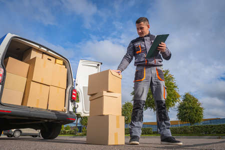 delivery service man. Delivery service worker next to van. Courier with large cardboard box. Courier on blue sky background. He provides courier service. Guy works in postal deliveryの写真素材