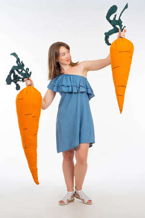 Giant carrot in hands of girl. Products in form of carrots. Girl is holding big pillows. Metaphor of growing carrots. Studio portrait of girl. She in denim dress. Woman on white backgroundの写真素材