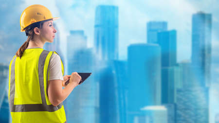 Engineer next to skyscrapers. Woman engineer. Girl in yellow uniform and helmet. Portrait of engineer in front of blurred skyscrapers. Construction company manager. Career in field constructionの写真素材