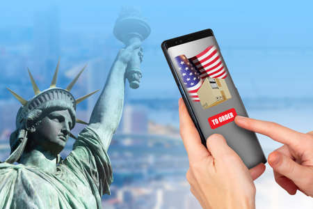 Ordering goods in USA. Purchase of American goods. Purchase from online store from America. Application with American flag on phone. Smartphone next to Statue of Liberty. USA online store applicationの写真素材