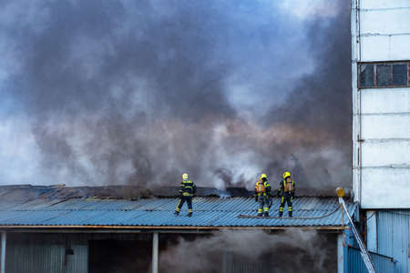 Fire at industrial facility. Fire service workers. Firefighters on roof of building. Extinguishing fire in factory. Several firefighters on background of smoke. Firefighters brigade during work.の写真素材