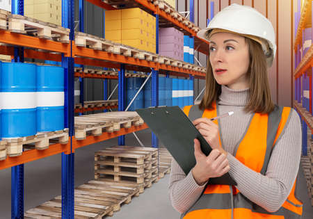 Chemical storage warehouse. Warehouse worker woman. Tanks chemical liquids behind her. Warehouse pallet system. Toxic barrels are stored on racks. Employee holds clipboard. Work in chemical industryの写真素材