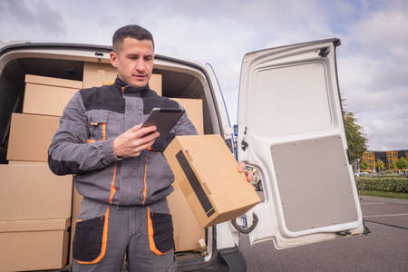Delivery man next to courier van. Postman looks into courier van. Cardboard boxes next to man. Courier van driver during work. Delivery service employee. Postal company worker holds clipboardの写真素材