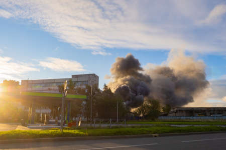 Smoke over enterprise. Smoke over industrial building. Smoke from fire against sky. Industrial factory on fire. Fume clubs in distance. Burning building on summer day. Fume over factory buildingの写真素材