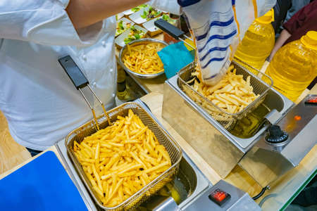 Cook fries french fries. A cook sprinkles french fries in a deep fryer. Sale of semi-finished products for a restaurant. Deep fryer during operation. Loaded deep fat fryer top view. Cook work.の写真素材