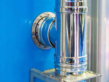 chimney pipe goes into wall. Shiny pipe on blue wall. Pipeline in production area. Stainless steel piping. Fragment of chimney system. Industrial engineering communications. Pipeline fragmentの写真素材