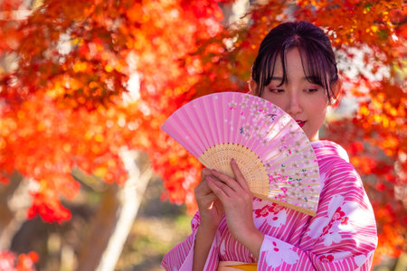 Japan. Geisha in the autumn park of Nara. Geisha on the background of red maple covers her face with a fan. Girl in japanese kimono poses with a fan. Japanese woman in pink kimono. 11.23.2019のeditorial素材