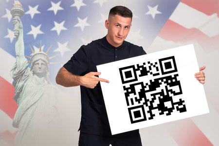 QR code in hands of man. American man demonstrates barcode. Guy in front of USA flag. Man points his...