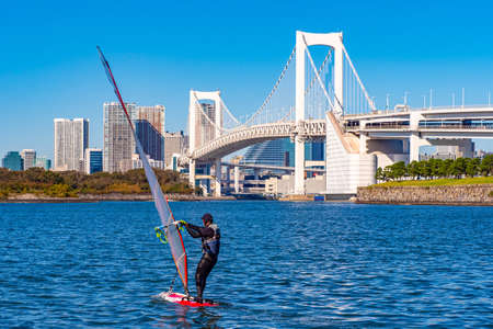 Japan. Windsurfer near Odaibo Island. Rainbow Bridge. View of Tokyo from Ostraibra Ostravo. Windsurfer in Tokyo Bay. Rainbow bridge on the background of blue sky. Japanese athlete.の写真素材