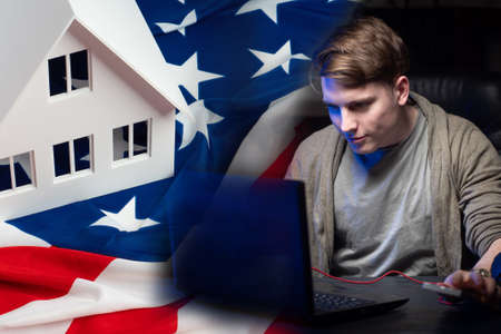 Home in the USA. Immigration to North America. A young guy next to a laptop. The layout of the house is on the flag. A man is looking for real estate on the Internet. Crossing. greencart lotteryの写真素材